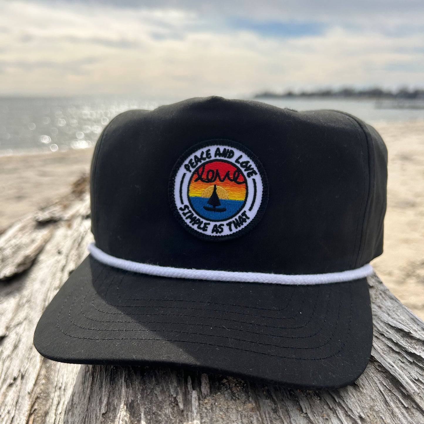 Black 'Peace and Love' rope hat from Simple As That, featuring a circular embroidered patch with a sunset, a peace sign, and the words 'Peace and Love' and 'Simple As That.' Accented with a white rope detail across the brim, displayed on driftwood by the beach with the ocean in the background.