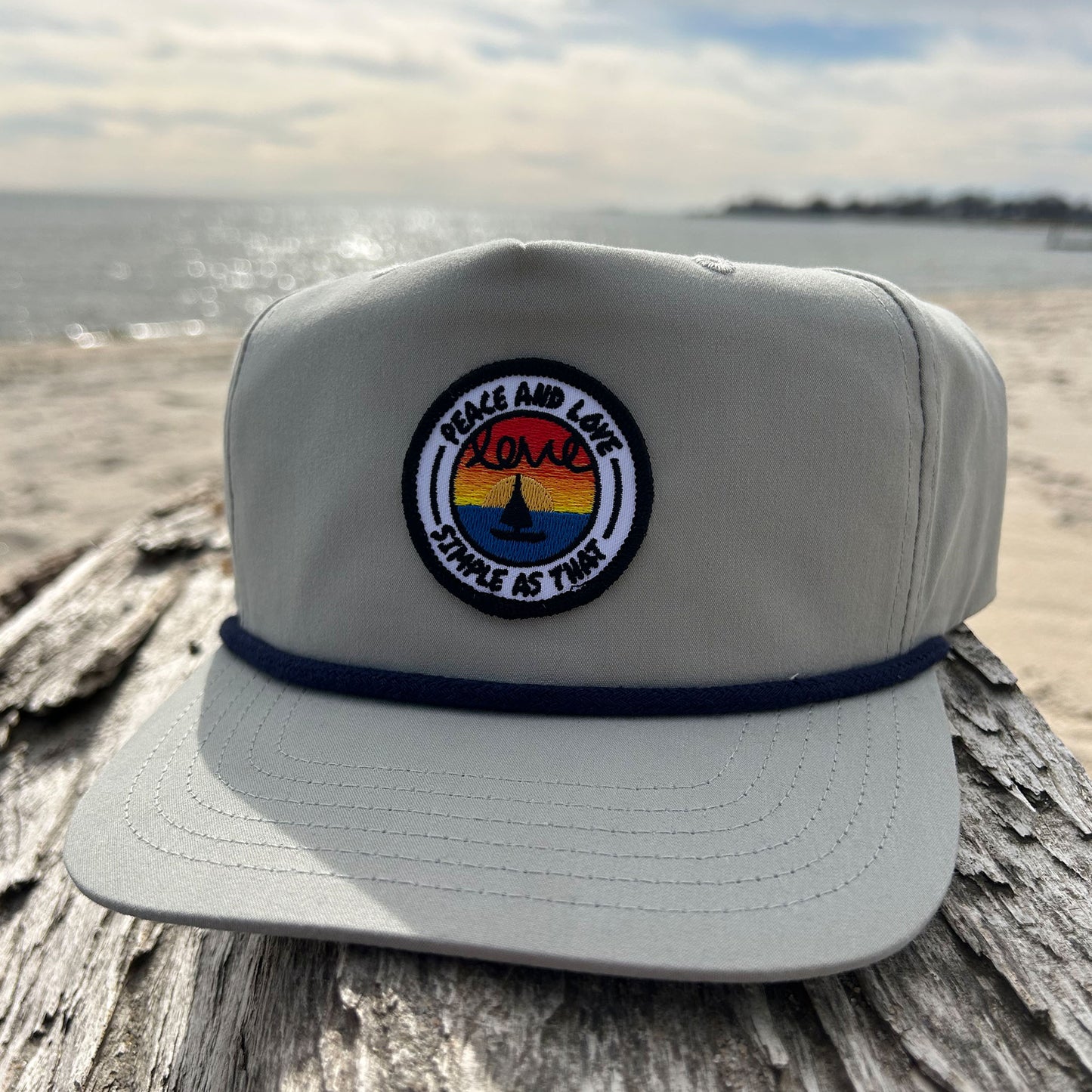 Grey 'Peace and Love' rope hat from Simple As That, featuring a circular embroidered patch with a sunset, a peace sign, and the words 'Peace and Love' and 'Simple As That.' Accented with a navy rope detail across the brim, displayed on driftwood by the beach with the ocean in the background.