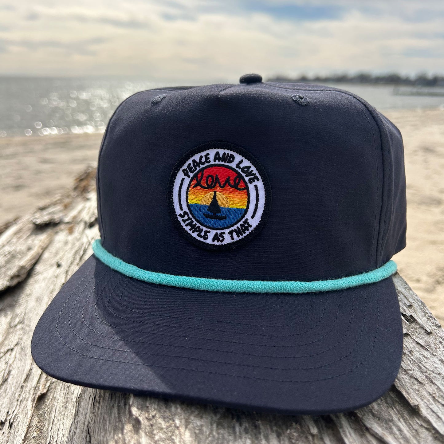 Navy blue 'Peace and Love' rope hat from Simple As That, featuring a circular embroidered patch with a sunset, a peace sign, and the words 'Peace and Love' and 'Simple As That.' Accented with a teal rope detail across the brim, displayed on driftwood by the beach with the ocean in the background.