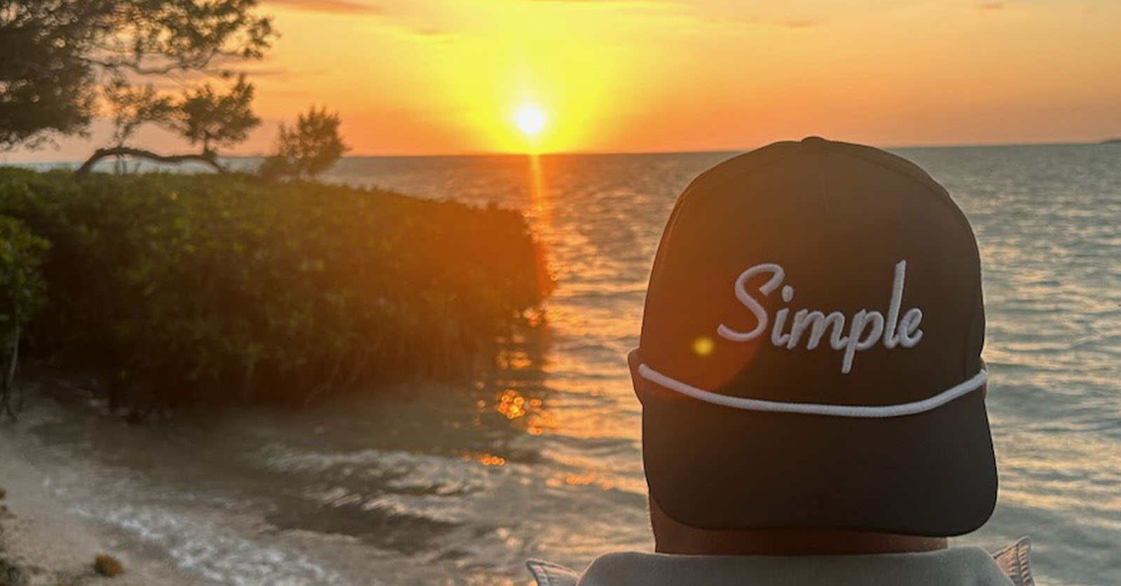 Person wearing a black 'Simple' rope hat while watching a stunning sunset over the ocean. The golden light reflects off the water, creating a peaceful and scenic moment that embodies the simplicity of life. It's really as Simple As That!