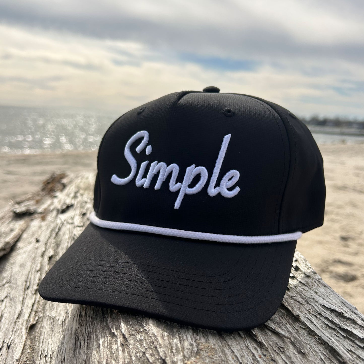 Black 'Simple' rope hat featuring bold white embroidered 'Simple' text on the front and a classic white rope detail across the brim. Displayed on driftwood at the beach with the ocean and sandy shore in the background, emphasizing its clean and minimalist design. It's really as Simple As That!