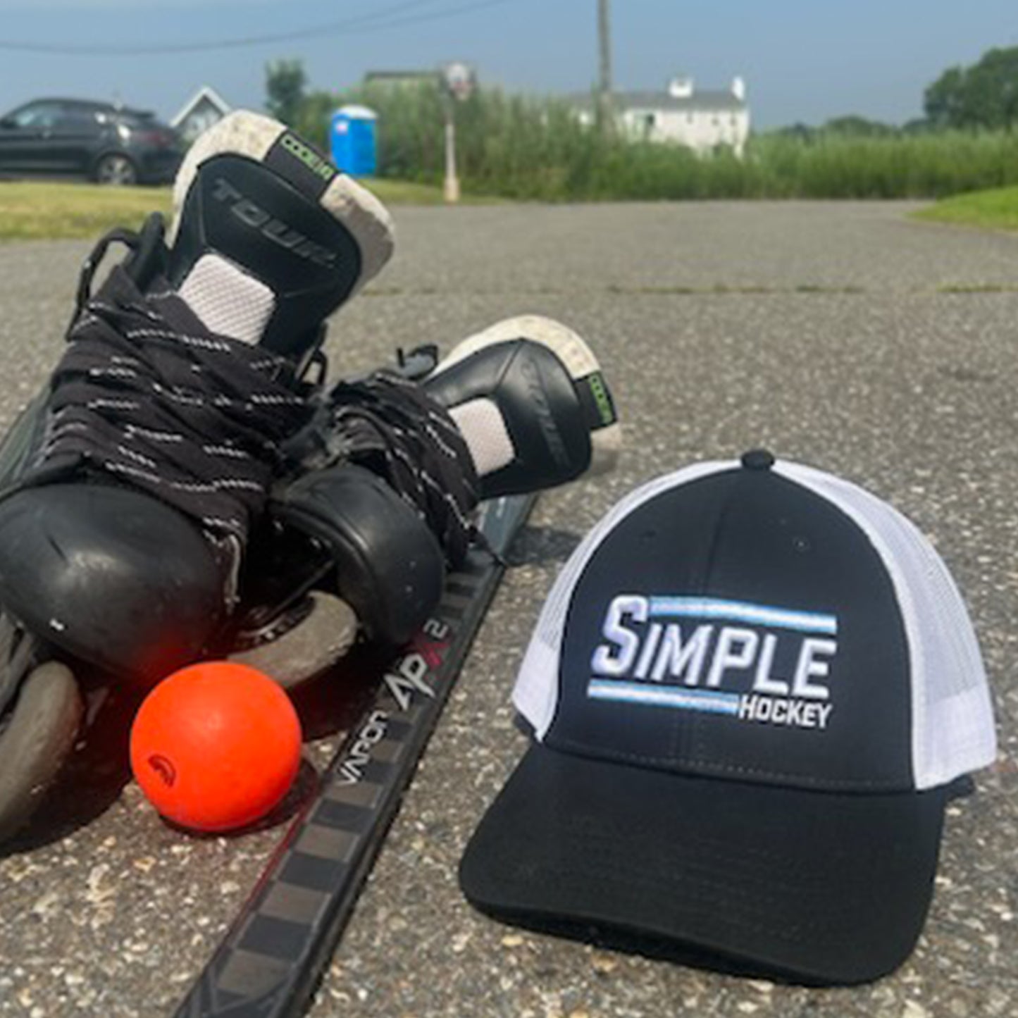 Black and white mesh-back 'Simple Hockey' hat displayed alongside roller hockey gear, including inline skates, a hockey stick, and an orange ball on a paved surface. Perfect for street hockey or on-ice play, this hat blends style and performance for hockey enthusiasts everywhere.