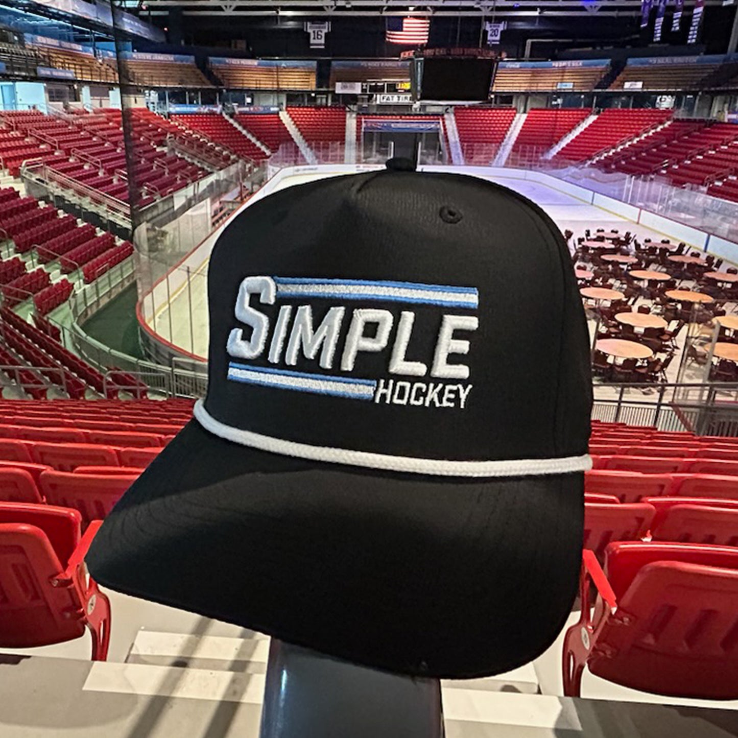 Black 'Simple Hockey' rope hat with bold white and light blue embroidered 'SIMPLE' text and 'HOCKEY' below, accented by two horizontal stripes and a white rope detail across the brim. The hat is displayed inside the iconic 1980 Herb Brooks Arena in Lake Placid, known for the 'Miracle on Ice, with red stadium seating and the ice rink in the background. It's really as Simple As That!