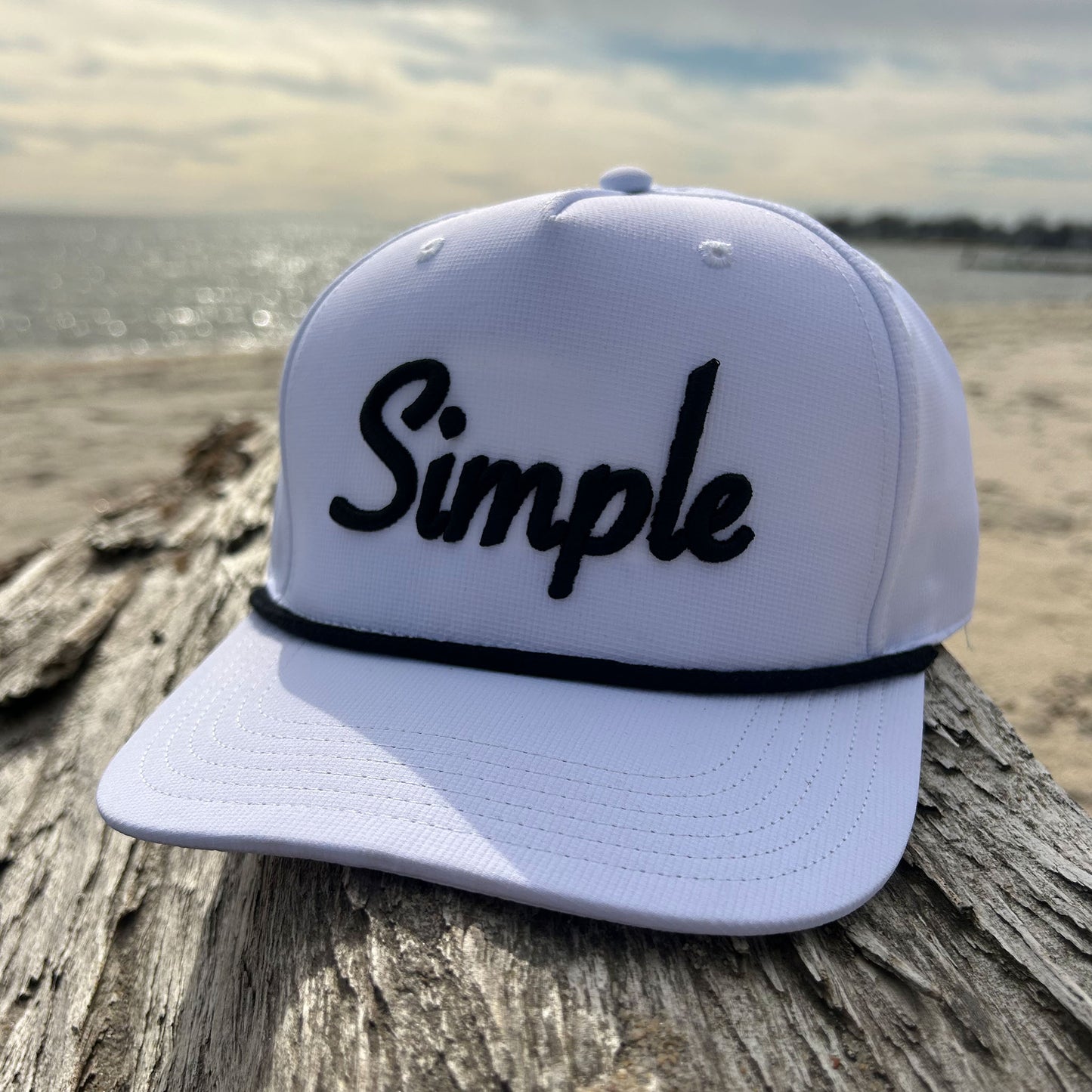 White 'Simple' rope hat featuring bold white embroidered 'Simple' text on the front and a classic white rope detail across the brim. Displayed on driftwood at the beach with the ocean and sandy shore in the background, emphasizing its clean and minimalist design. It's really as Simple As That!