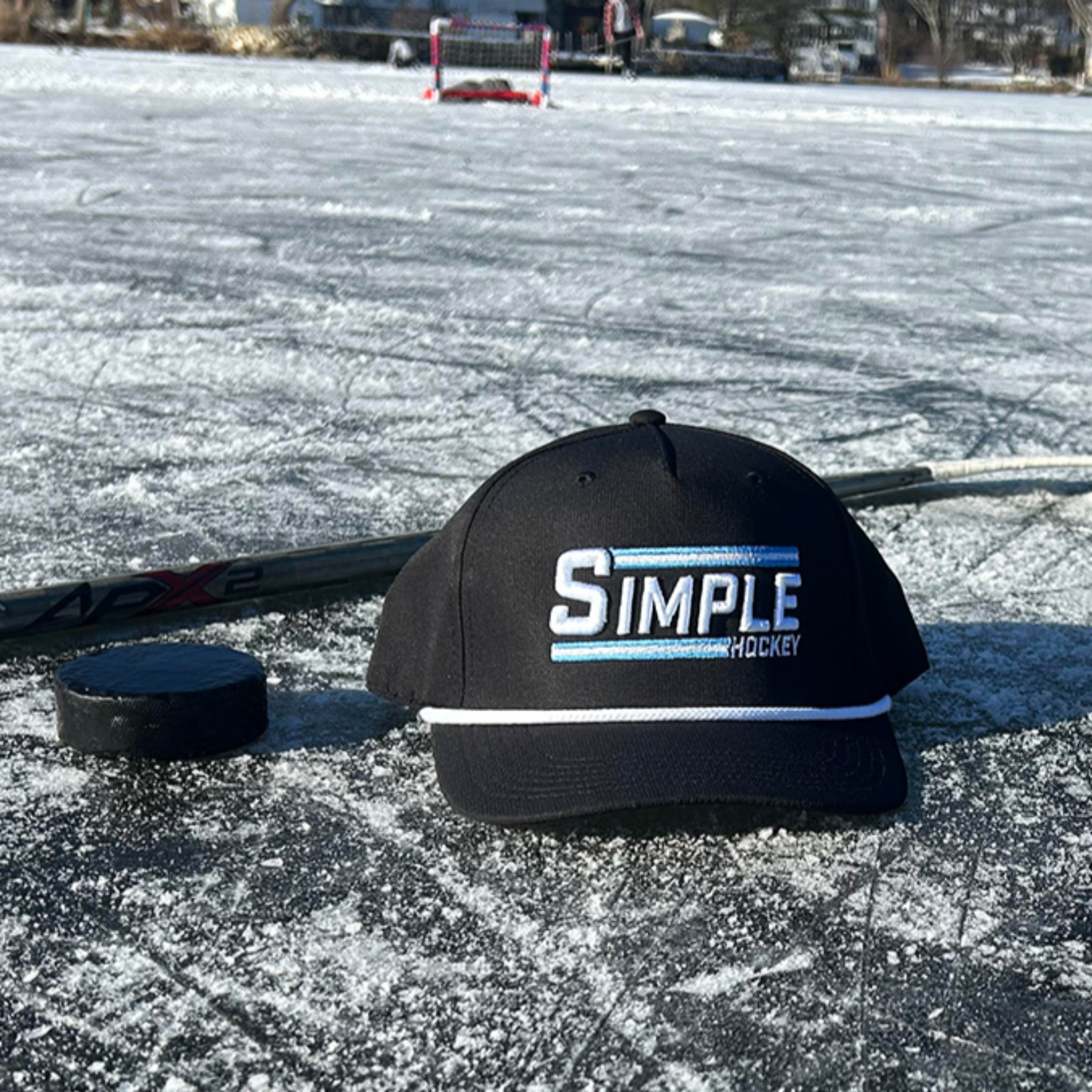 Simple Hockey rope hat placed on a frozen outdoor rink alongside a hockey puck, stick, and gloves. The sun shines on the ice, capturing the essence of outdoor pond hockey with a crisp winter backdrop. It's really as Simple As That!