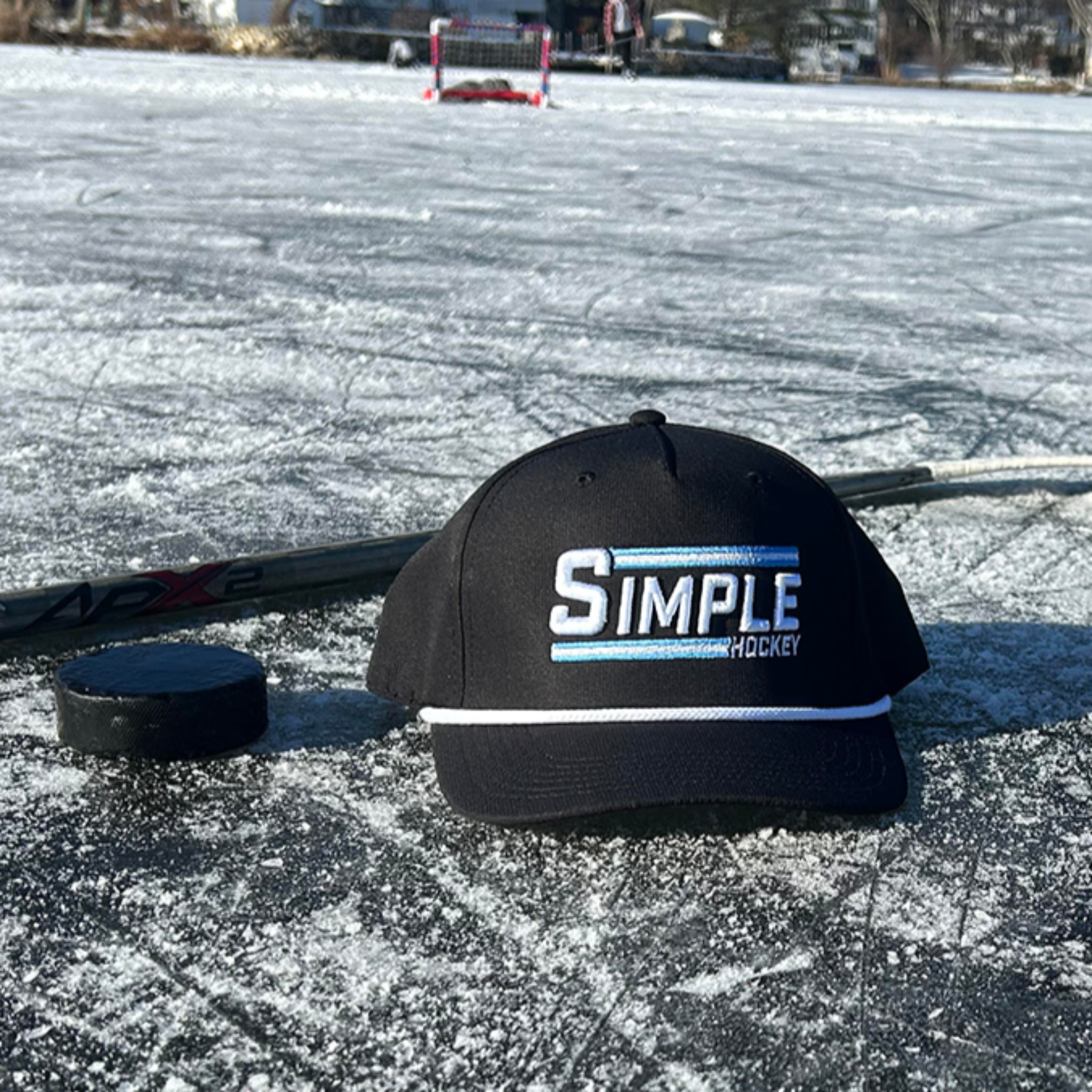 Simple Hockey rope hat placed on a frozen outdoor rink alongside a hockey puck, stick, and gloves. The sun shines on the ice, capturing the essence of outdoor pond hockey with a crisp winter backdrop. It's really as Simple As That!
