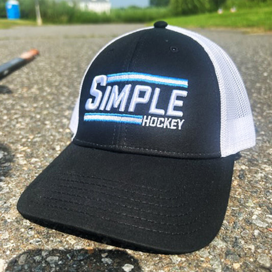 Black and white mesh-back 'Simple Hockey' hat featuring bold white and light blue embroidered 'SIMPLE' text with 'HOCKEY' below. The front panel is solid black with a curved brim, while the breathable white mesh back offers comfort and ventilation. Displayed outdoors on a paved surface, perfect for hockey enthusiasts on and off the rink.