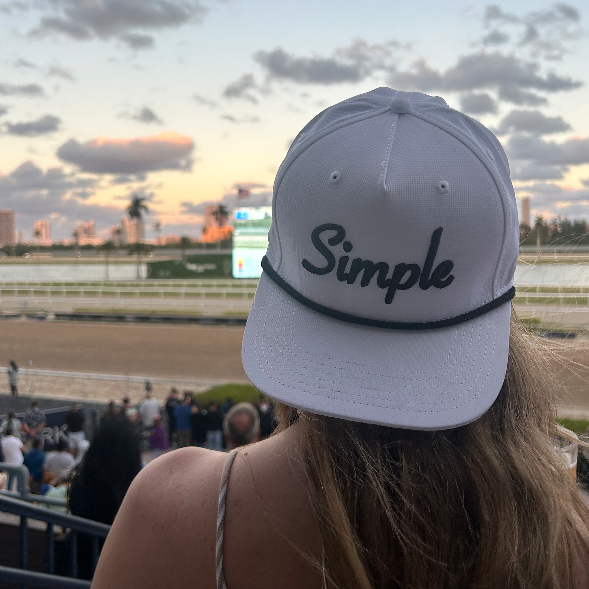 An individual wearing a white Simple rope hat while watching a horse race at sunset. The sky is filled with soft clouds, and the city skyline is visible in the background, creating a relaxed and stylish atmosphere. It's really as Simple As That!